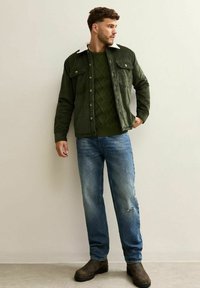 Chaqueta de pana verde con un cuello blanco, superpuesta a un suéter verde texturizado, combinada con jeans azules desgastados y botas marrones.
