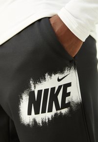 Mustat urheiluhousut, joissa on suuri valkoinen Nike-logo suihkumaalitekstuurilla vasemmassa alakulmassa, on valmistettu sileästä kankaasta.
