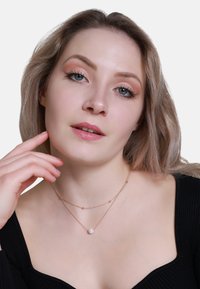 Collier superposé avec une délicate chaîne en or rose ornée de petites perles et d'une perle blanche centrale, porté sur un haut noir côtelé.