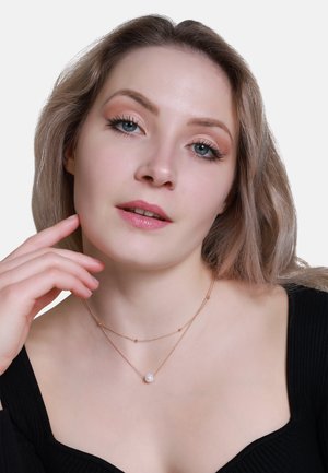 Gestaffelte Halskette mit einer zarten roségoldenen Kette, kleinen Perlen und einer zentralen weißen Perle, getragen über einem schwarzen gerippten Oberteil.