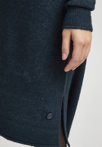 Robe tricotée bleu marine foncé avec des poignets côtelés, présentant une fente sur le côté et un accent de bouton bleu marine à l'ourlet. Texture douce et coupe relaxée.