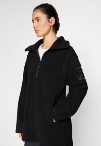 Pullover in pile nero con colletto a mezza zip, esterno texturizzato, due tasche laterali e dettaglio a toppa sulla manica.