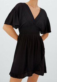 Nainen, joka wear short black wrap dress with wide flutter sleeves, smocked waist, and ruffled hem vaaleataustalla.