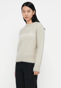 Lichtgrijze, fuzzy trui met geribde ronde hals en verlaagde schouders. Bevat het "Calvin Klein" logo in een lichtere tint. Gestyled met zwarte broek.