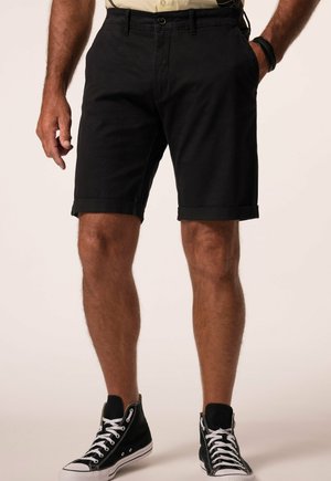 Mand iført sorte op-rullede shorts, sorte high-top sneakers med hvide snørebånd og en lysefarvet skjorte, står med den ene hånd i lommen.