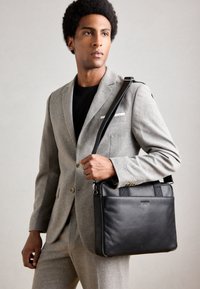 Ungdommelig mann i lys grå dress som bærer en svart skinn skulderveske, som ser litt til siden mot en enkel bakgrunn.