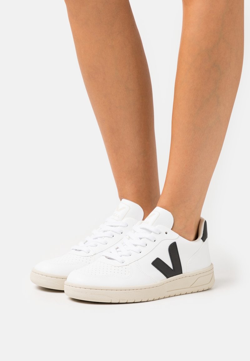 Jambes portant des baskets blanches avec des lacets blancs, des semelles beige et un logo noir "V" sur le côté sur fond blanc.