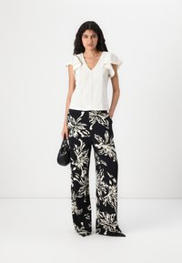Top alb cu model de ochi, cu mâneci volan, purtat cu pantaloni negri largi, cu un imprimeu floral alb îndrăzneț; ținând o geantă neagră.