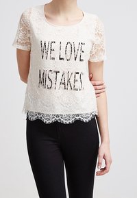 Top de manga corta de encaje blanco con la inscripción "AMAMOS LOS ERRORES" impresa en negro. El dobladillo de encaje con forma de olas añade detalle; combinado con pantalones negros.