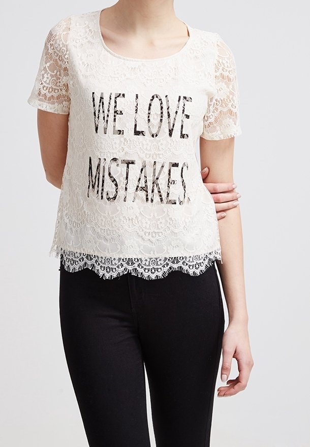 Top de manga corta de encaje blanco con la inscripción "AMAMOS LOS ERRORES" impresa en negro. El dobladillo de encaje con forma de olas añade detalle; combinado con pantalones negros.
