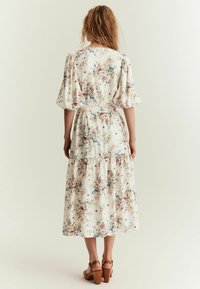 Blomster midi kjole i cremet stof. Har pufærmer, en taljebånd, en lagdelt nederdel med flæser og en blanding af pink og blå blomsterprint.