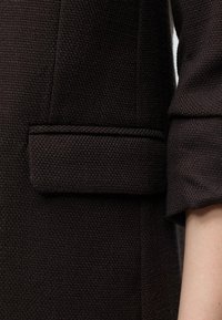 Gros plan d'une veste en tissu noir texturé avec une poche à rabat et une partie d'une manche retroussée montrant une peau claire.