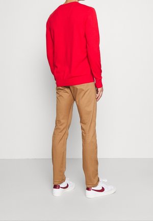 Persoon draagt een felrode trui met lange mouwen, beige broek en witte Nike-sneakers met een rode swoosh, staand tegen een witte achtergrond.