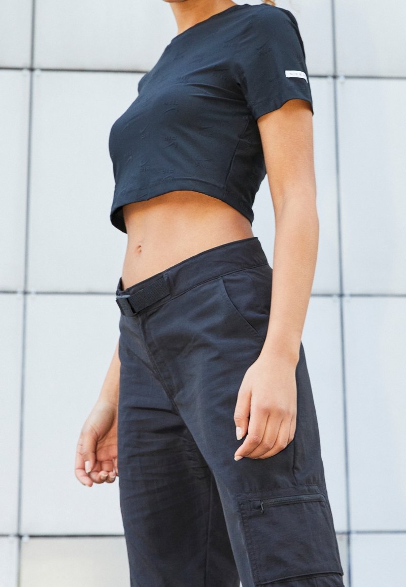 Personne portant un haut court noir à manches courtes et un pantalon cargo noir avec ceinture, debout contre un mur carrelé clair.