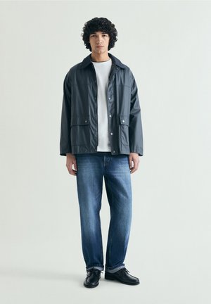 Jeune homme aux cheveux bouclés portant une veste noire boutonnée, un t-shirt blanc, un jean bleu et des chaussures noires, se tenant devant un fond clair uni.