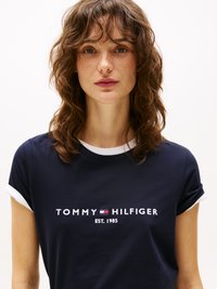 Jonge vrouw met golvend bruin haar en sproeten, gekleed in een marineblauw Tommy Hilfiger T-shirt met opgerolde witte mouwen.