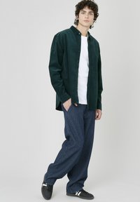 Chemise en velours côtelé vert foncé sur un t-shirt blanc, associée à un pantalon large à rayures bleues et des baskets noires. Design simple avec des boutons.