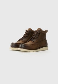 Botas de cuero marrón hasta el tobillo con punteras redondas, suelas blancas que contrastan y cordones oscuros. Presentan un diseño cosido y un pequeño tirador en el talón.