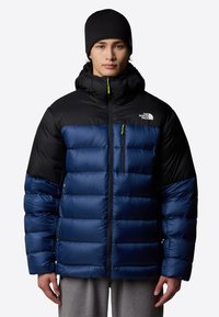 Niebieska i czarna kurtka puchowa z kapturem, z kwadratowymi przeszyciami, z przodu zapinana na zamek oraz z kieszenią na piersi. Odbijające światło logo na klatce piersiowej.