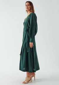Green long-sleeve dress with a deep V-neck, cinched waist tie, and pleated skirt. Worn with beige high-heeled sandals.
