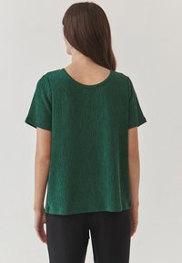 Groen textuur met korte mouwen top met een ronde halslijn. Heeft een losse pasvorm en subtiele verticale lijnen, gecombineerd met zwarte broeken.