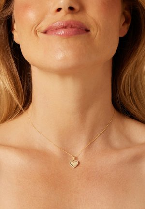 Collier en or avec un pendentif en forme de cœur bordé de petites pierres claires, reposant sur un cou nu. Le pendentif présente une surface intérieure lisse.