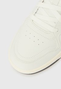 Close-up do bico do ténis branco com painéis costurados, orifícios de ventilação e atacadores planos presos, sobre um fundo claro.