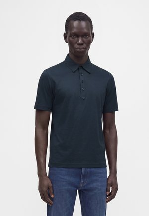 Mann trägt ein dunkles kurzärmeliges Poloshirt mit Knöpfen und blaue Jeans, steht vor einem schlichten hellen Hintergrund.