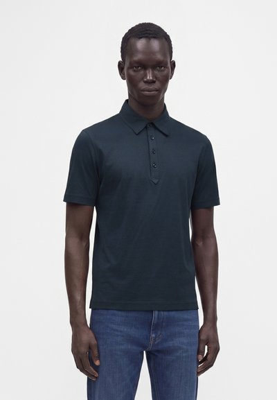 Homme portant un polo sombre à manches courtes avec des boutons et un jean bleu, debout devant un fond clair uni.