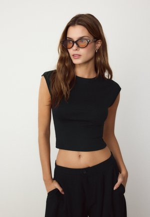 Junge Frau mit langen braunen Haaren, trägt ein schwarzes ärmelloses Crop-Top, schwarze Hose und dunkle Sonnenbrille, steht vor einem einfarbigen Hintergrund.