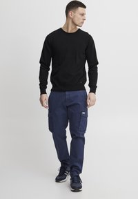 Svart strikkegenser, marineblå cargo-bukser med sidelommer, og mørke joggesko. Modell går foran en ensfarget bakgrunn.