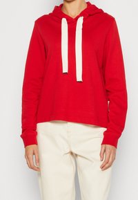 Sudadera corta roja con mangas largas, cordones blancos y puños de canalé. La tela parece suave con una textura lisa. Etiqueta blanca en el lateral.