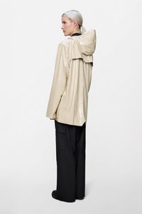 Μπεζ γυαλιστερό αδιάβροχο με κουκούλα, με φαρδιά εφαρμογή και πτερύγιο στην πλάτη, σε συνδυασμό με μαύρο παντελόνι φαρδιάς γραμμής και μαύρα υποδήματα.