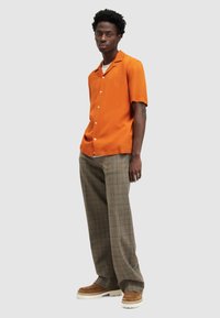 Camisa de botones naranja de manga corta combinada con pantalones marrón de cuadros y zapatos sin cordones color marrón claro, mostrando un atuendo relajado pero coordinado.