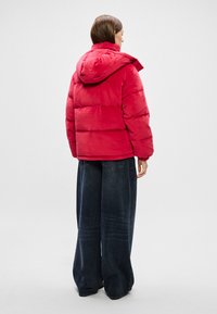 Sarkans pūkains džemperis ar kapuci, ar izšūtu dizainu un nedaudz spīdīgu tekstūru. Apvienots ar platām kājām tumšām džinsu biksēm.