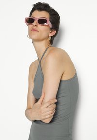 Top de alças finas cinza com texturização, combinado com óculos de sol retangulares rosa com lentes escuras. Modelo com cabelo curto e brincos de argola dourados.