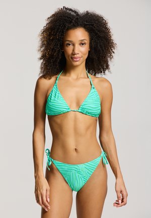 Žena s kučeravými vlasmi, ktorá má na sebe zelené pruhované bikiny s halter topom a spodným dielom so zaväzovaním na stranách, stojaca pred jednofarebným svetlým pozadím.