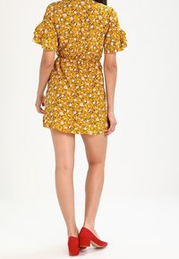 Gul blommig klänning med korta volangärmar, samlad midja och utställd nederkant. Matchad med röda klackar med kilklack. Bakvy visas.