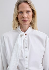 Blusa blanca de corte holgado, con un lazo de tela en el cuello y botones plateados decorativos. Textura suave con un cuello clásico.