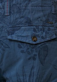 Blue fabric with a floral pattern features a patch pocket with a brown button. Includes contrasting red stitching and a label on the side.