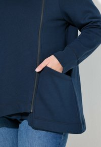 Camiseta de manga larga en color navy, hecha de una tela suave, con cierre de cremallera y un bolsillo frontal, además de detalles de costura visibles.