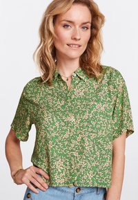 Kurzärmlige grüne Bluse mit einem floralem Muster in Beige, ausgestattet mit einem durchgehenden Knopfverschluss und einem lockeren Schnitt. Sanfter Stoff mit leichter Textur.