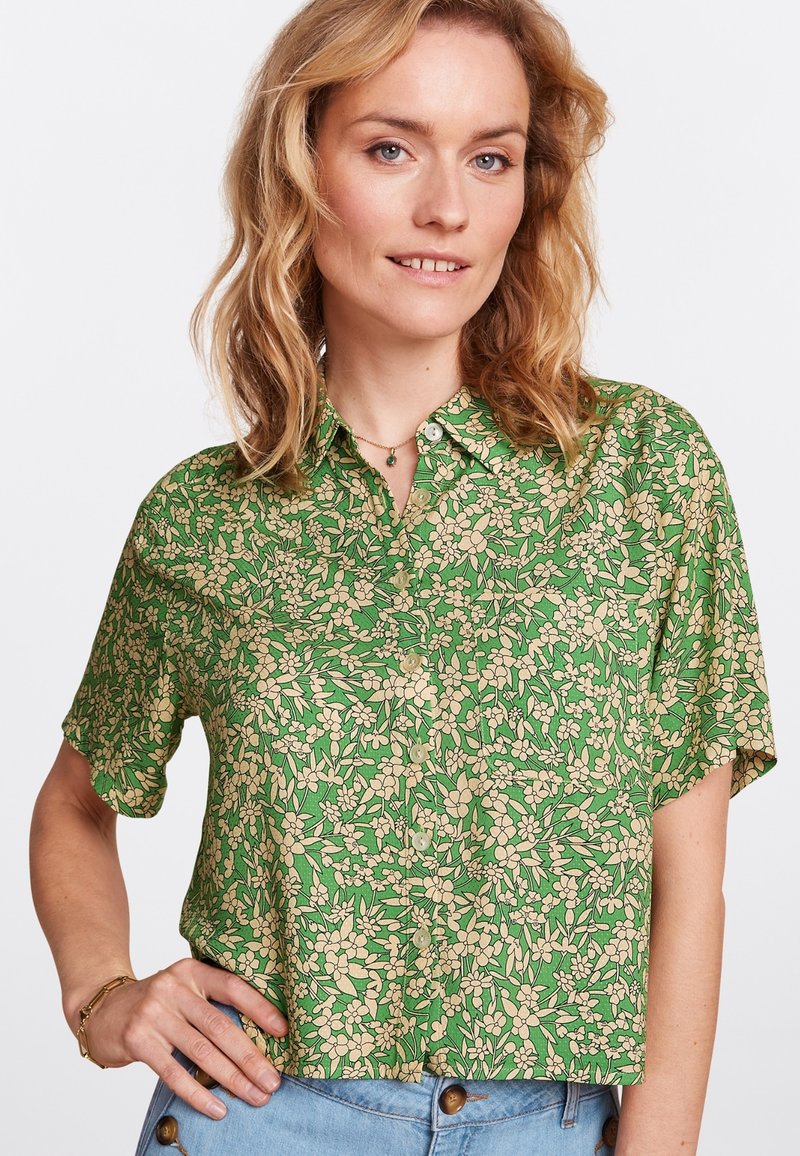 Kurzärmlige grüne Bluse mit einem floralem Muster in Beige, ausgestattet mit einem durchgehenden Knopfverschluss und einem lockeren Schnitt. Sanfter Stoff mit leichter Textur.