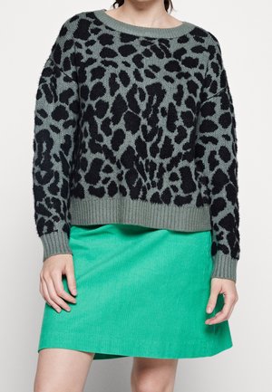 Femme portant un pull gris à imprimé léopard noir et une jupe en velours côtelé vert vif, debout devant un fond uni.