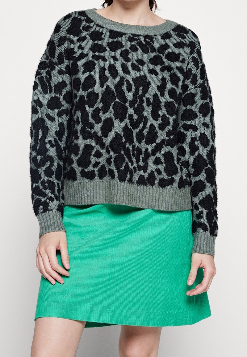 Femme portant un pull gris à imprimé léopard noir et une jupe en velours côtelé vert vif, debout devant un fond uni.