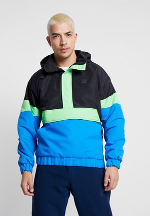 Huva-pulloverjacka med svarta, gröna och blå paneler, elastiska ärmslut och en framhalvsdragkedja. Tillverkad av lätt, slätt material.