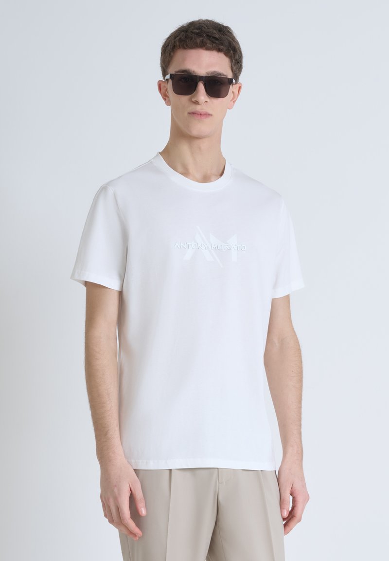 Camiseta de algodón blanca con cuello redondo y mangas cortas, que presenta un sutil logo en relieve en el pecho. La tela es suave y ligera.
