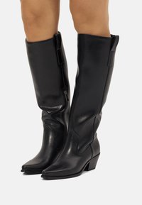 Botas de cuero negro hasta la rodilla con punta afilada, cremallera lateral y detalles en costura angular, con un tacón bajo tipo bloque. Textura suave.