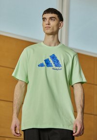 T-shirt oversize verde chiaro in cotone, con un design grafico blu di tre strisce e la parola "Basketball" sottostante.