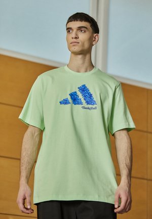 T-shirt oversize verde chiaro in cotone, con un design grafico blu di tre strisce e la parola "Basketball" sottostante.